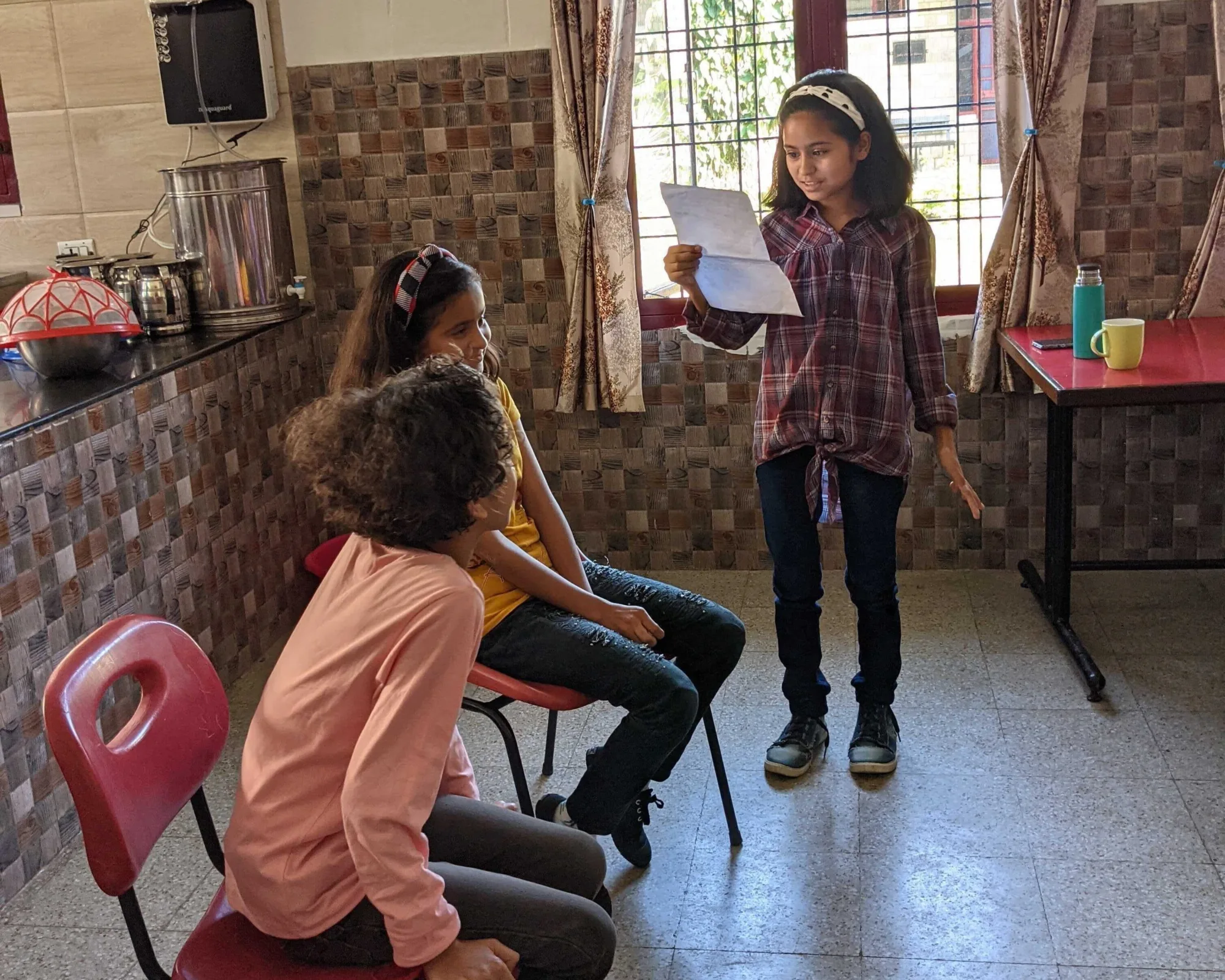 One young girl standing reading a text to two seated girls at Pestalozzi International India.