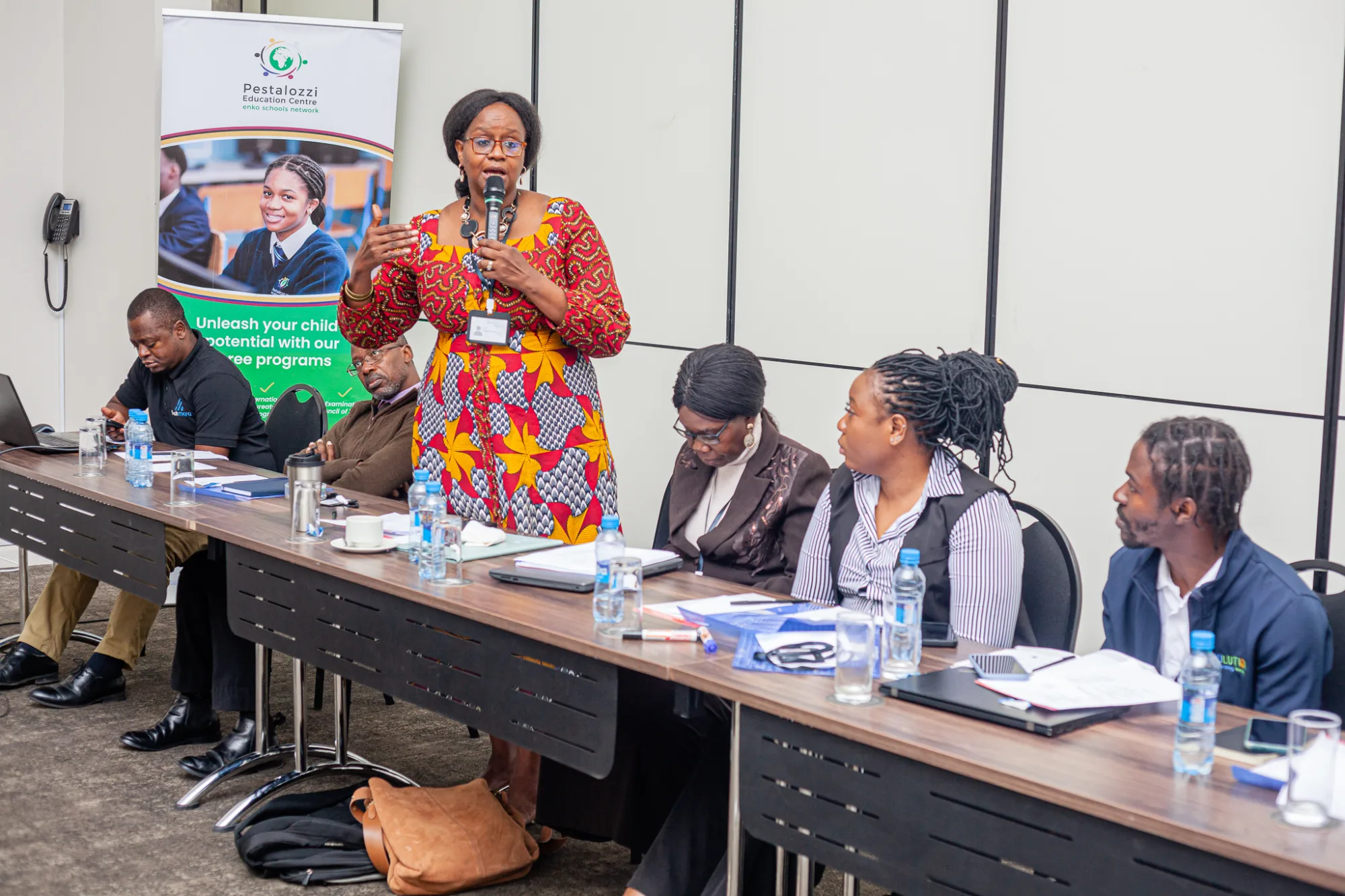 Image shows Pestalozzi Zambia's CEO at Zambia's roundtable event in August 2024standing and sharing her ideas with staff, alumni and businesses around the new SkillsHub Programme. in the image 5 individuals can be seen seated listening intently.